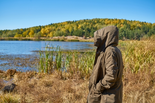 茂盛树林旁的湖泊边一名穿着大衣带着兜帽的男子